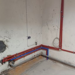 AGAMAR - Instalacja rur: czerwone i niebieskie rury prowadzone wzdłuż ściany, mocowane klamrami. Widoczne także białe rury i szare rury kanalizacyjne. Surowe wykończenie ścian.