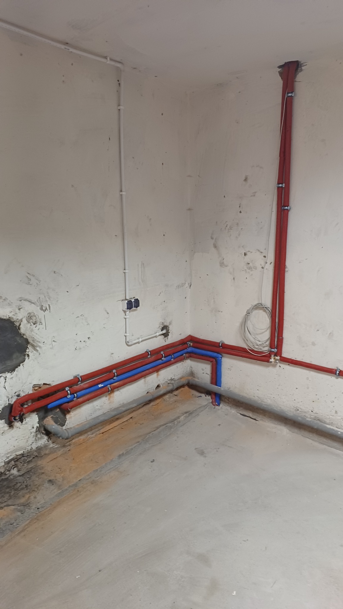 Instalacja rur: czerwone i niebieskie rury prowadzone wzdłuż ściany, mocowane klamrami. Widoczne także białe rury i szara rura kanalizacyjna przy podłodze. Surowe wykończenie.