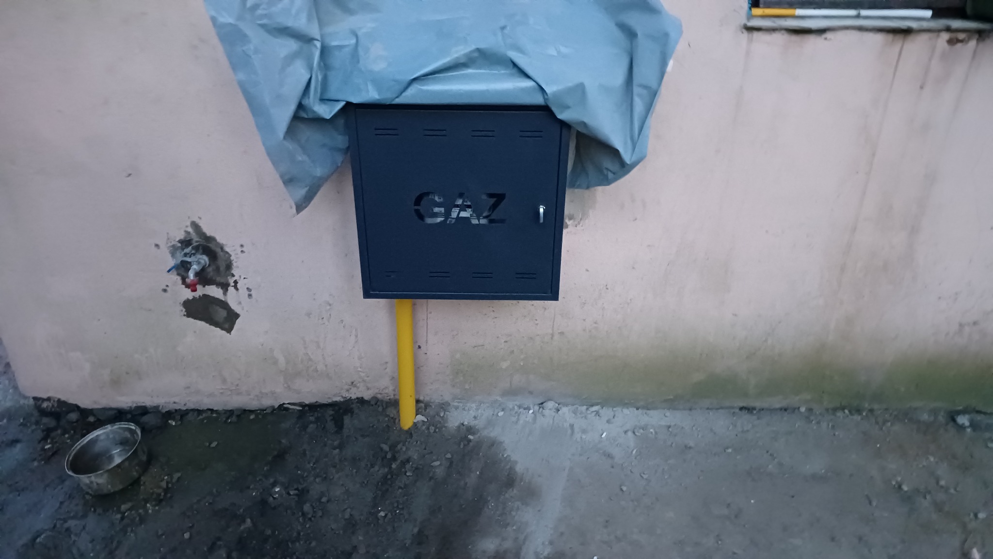 Zamontowana, czarna skrzynka gazowa z napisem 'GAZ' na elewacji budynku, z widoczną żółtą rurą doprowadzającą gaz. Częściowo przykryta folią ochronną.