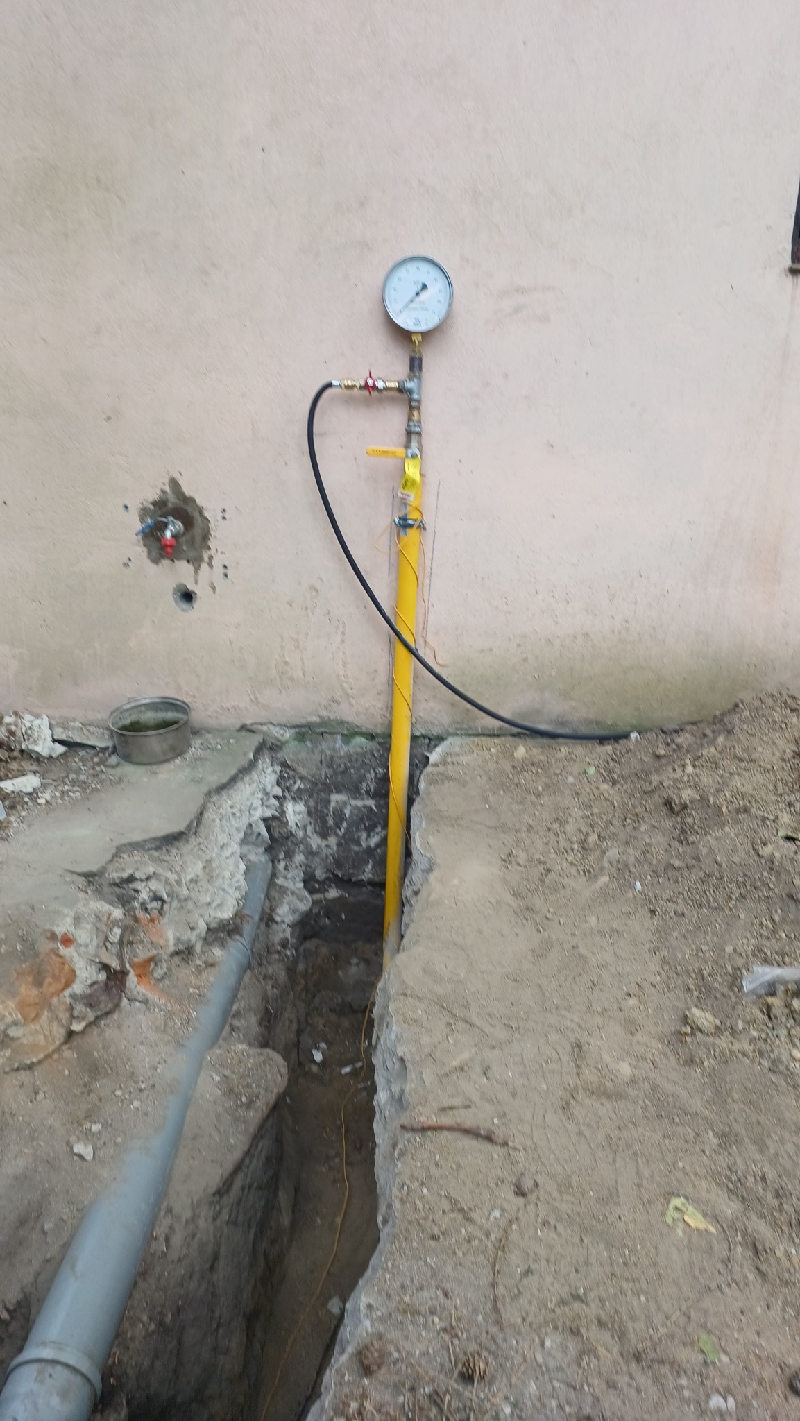 Żółta rura gazowa z manometrem wystająca z wykopu przy ścianie budynku. Widoczne przyłącza i fragment rury kanalizacyjnej. Prace ziemne w toku.