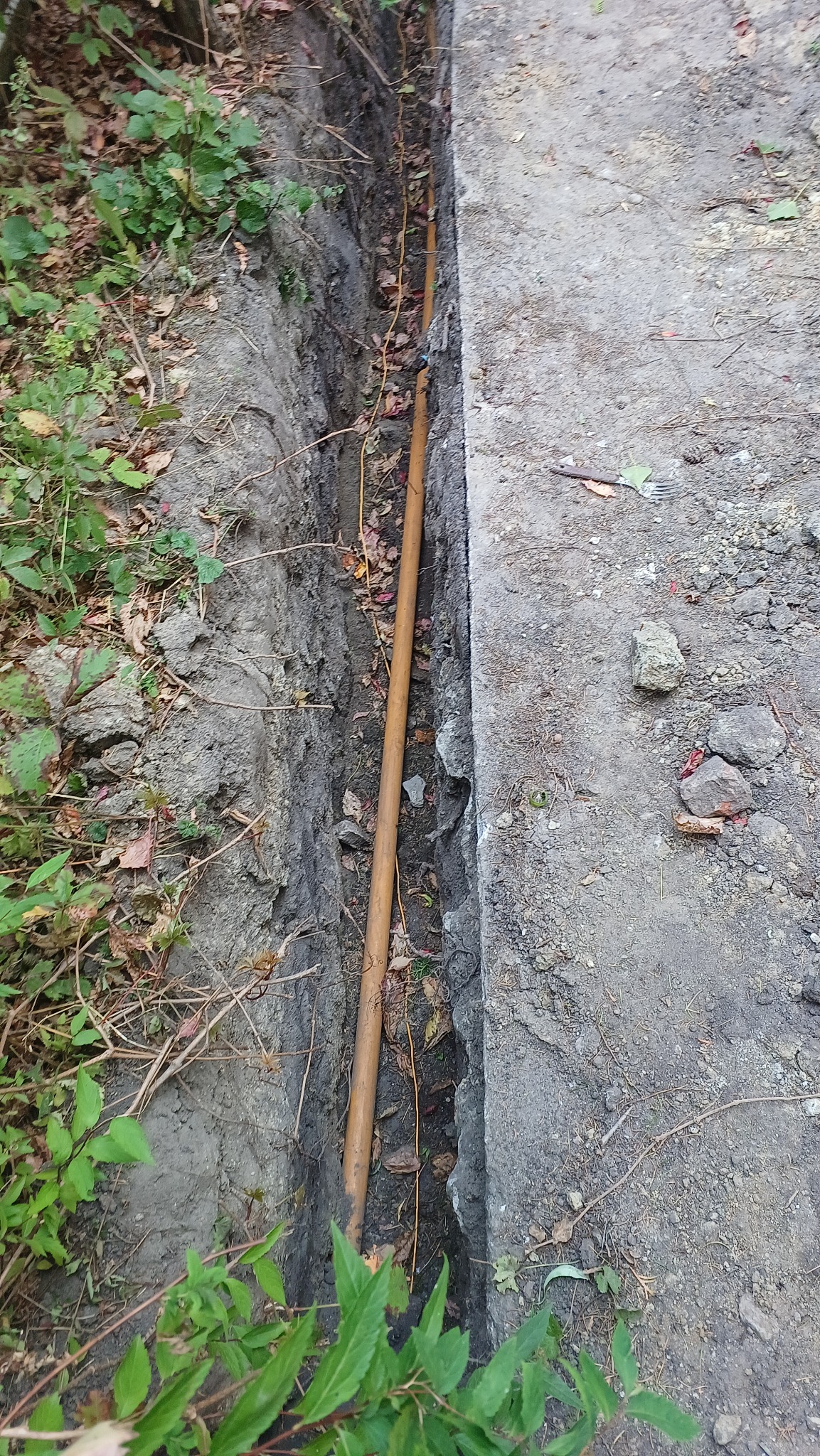 Rura w wykopie ziemnym, prawdopodobnie instalacja wodno-kanalizacyjna lub gazowa, widok z góry, obok betonowa płyta chodnikowa.