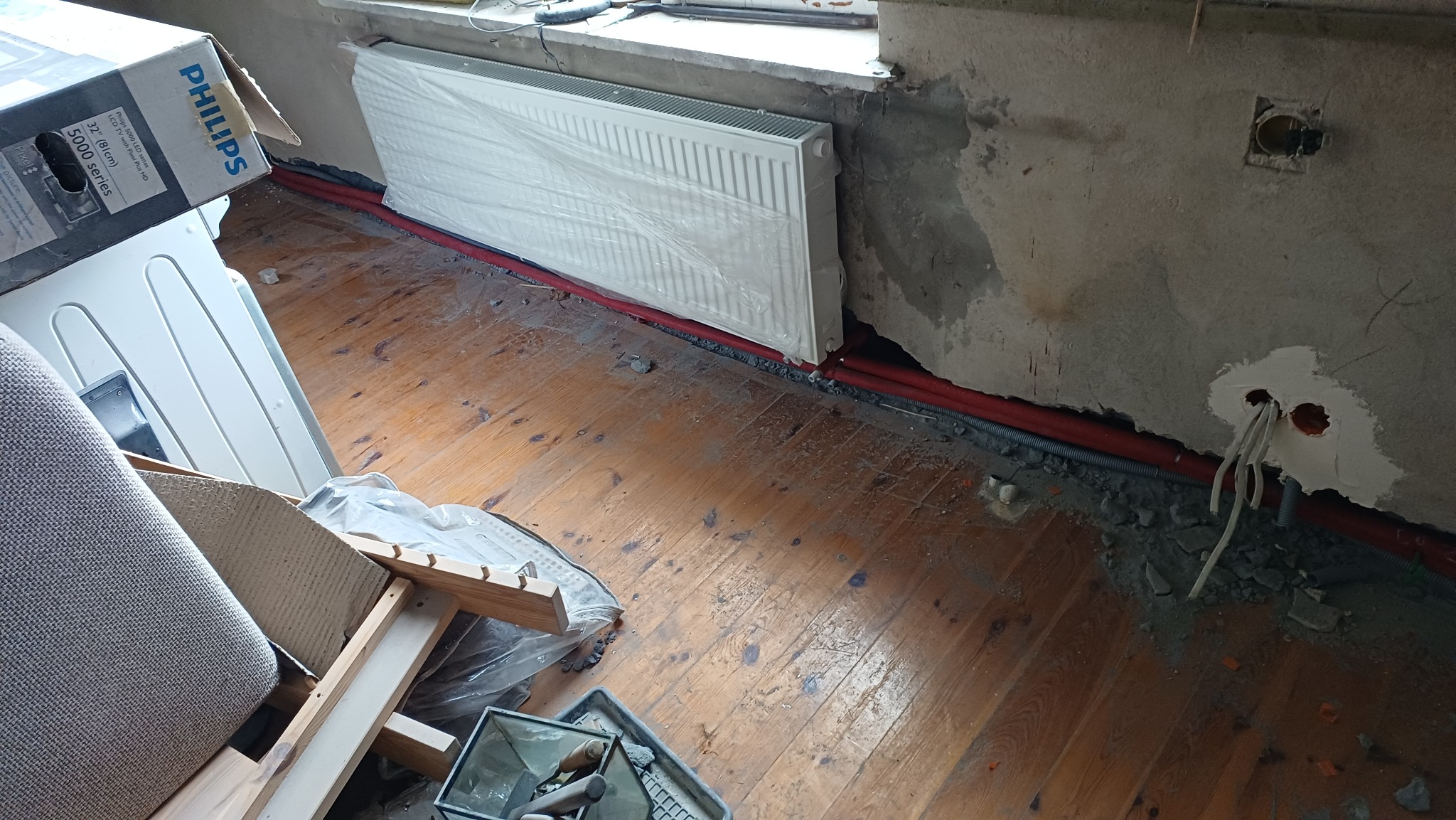 Montaż grzejnika panelowego, zabezpieczonego folią, z widocznymi czerwonymi rurami instalacji grzewczej wzdłuż ściany. Surowe wykończenie ścian, drewniana podłoga.