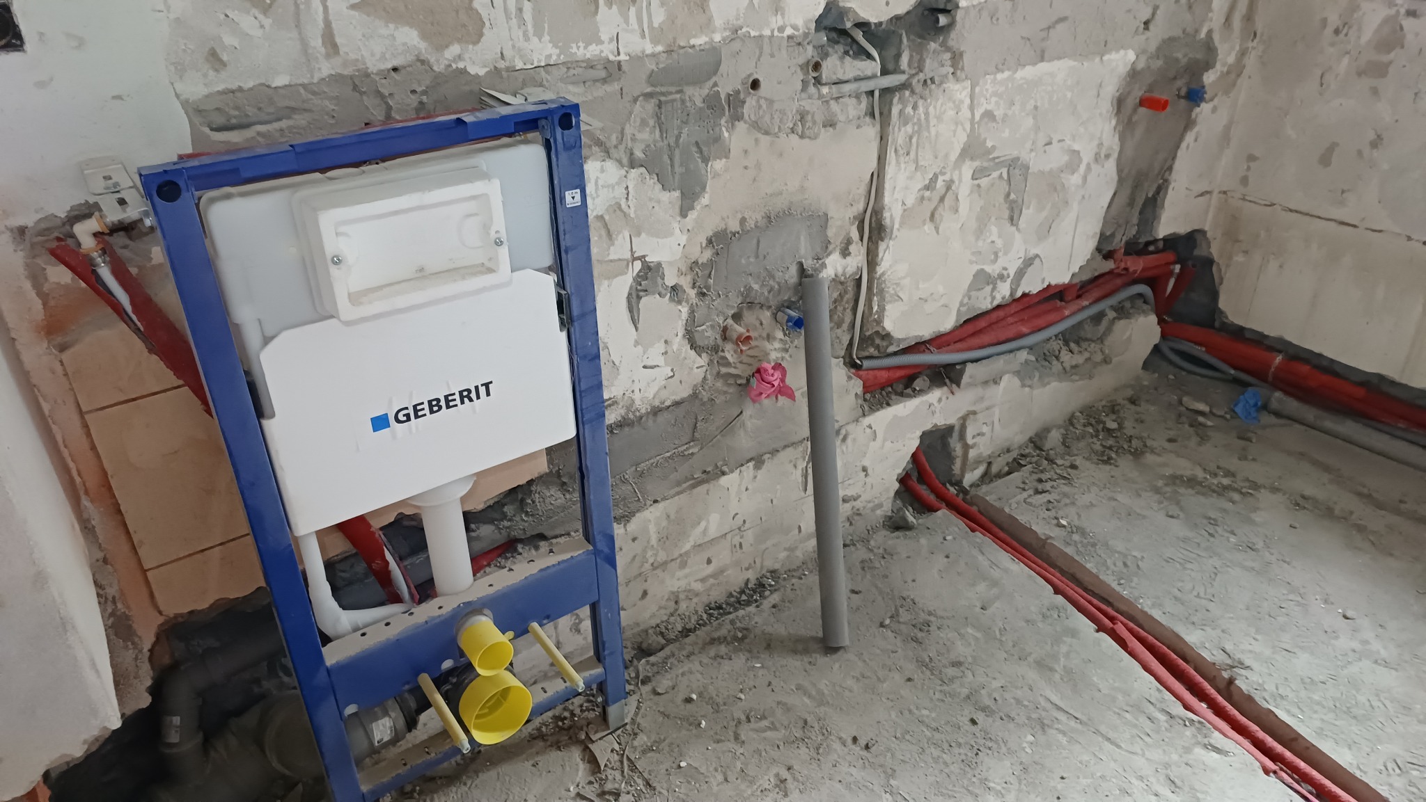 Stelaż podtynkowy Geberit w surowym wnętrzu łazienki, widoczne czerwone rury instalacji wodnej w ścianie i na podłodze, szary pion kanalizacyjny. Etap prac instalacyjnych.