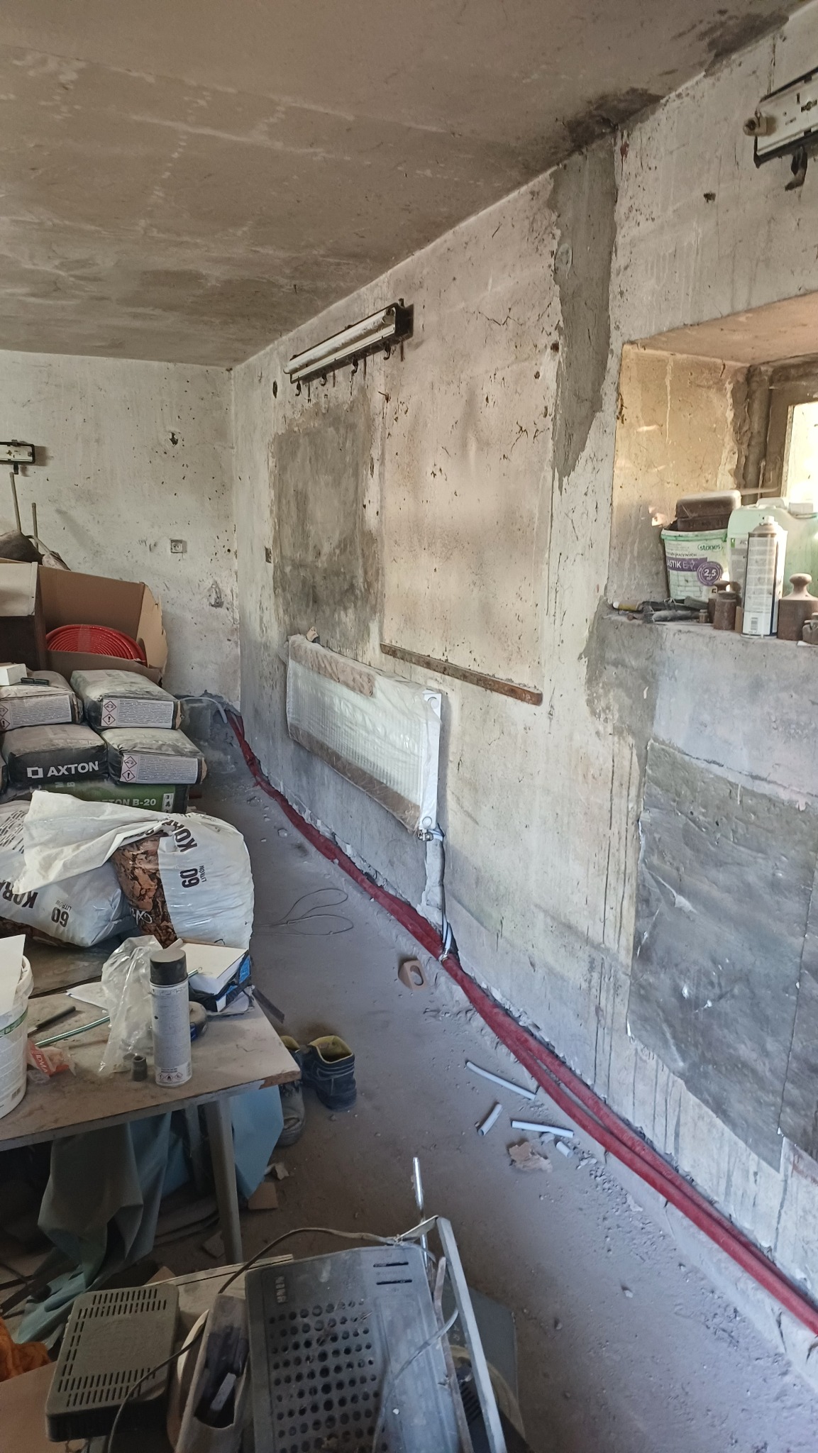 Wnętrze w trakcie remontu: grzejnik osłonięty folią, czerwone rury instalacji grzewczej wzdłuż ściany, worki z zaprawą i narzędzia. Surowy beton i widoczne ślady prac.