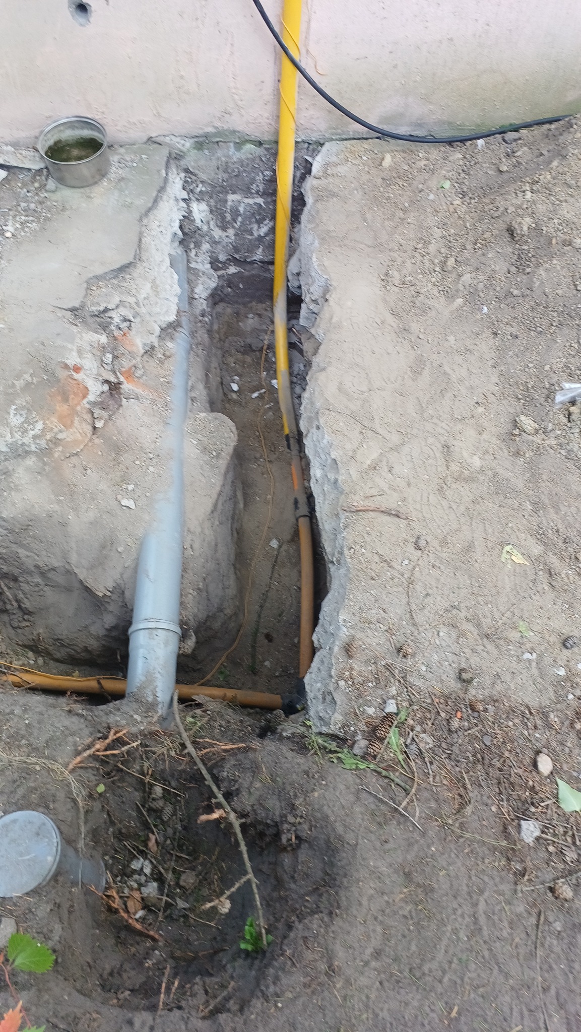 Wykop z widocznymi rurami: szara pionowa, żółta pionowa i brązowa pozioma. Fragment fundamentu i ziemia. Widoczne połączenia rur. Prace ziemne w toku.