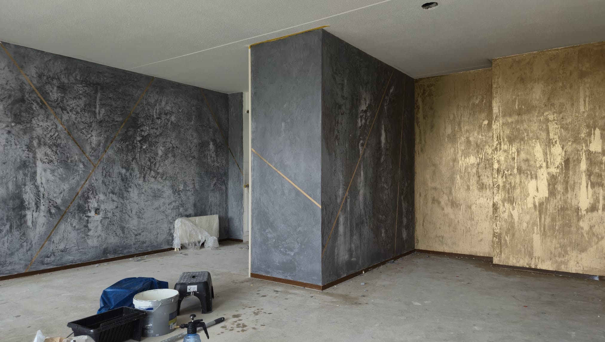 Pokój po dekoracyjnym malowaniu ścian w odcieniach szarości i złota, z geometrycznymi wzorami. Widoczne narzędzia malarskie na podłodze, co sugeruje świeżo zakończone prace.