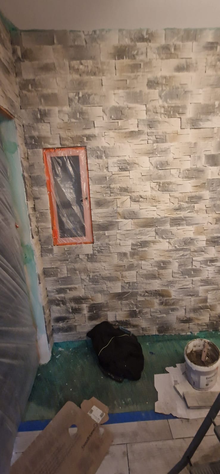 Ściana wykończona dekoracyjną, szarą okładziną kamienną. Okno zabezpieczone folią. Na podłodze wiadro z zaprawą i karton. Widoczne ślady prac wykończeniowych.