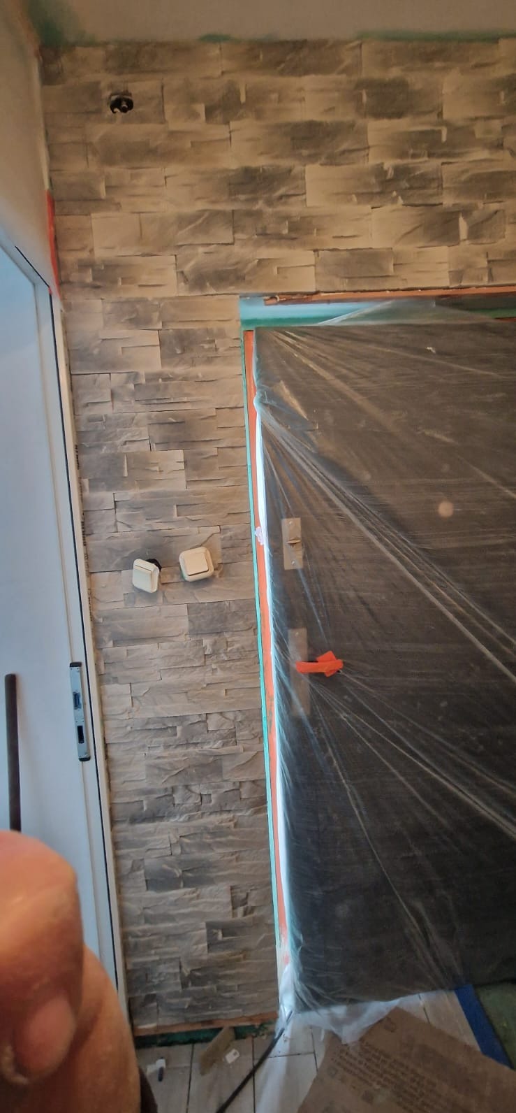 Ściana wykończona dekoracyjną okładziną imitującą kamień, z widocznymi włącznikami światła. Drzwi zabezpieczone folią ochronną podczas prac wykończeniowych.