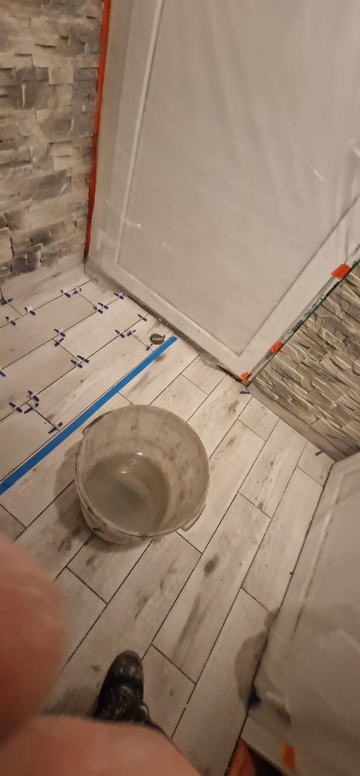 Ułożone płytki podłogowe w łazience z krzyżykami dystansowymi i wiaderkiem z wodą. Fragment ściany wykończony imitacją kamienia. Zabezpieczona folią strefa prysznica.
