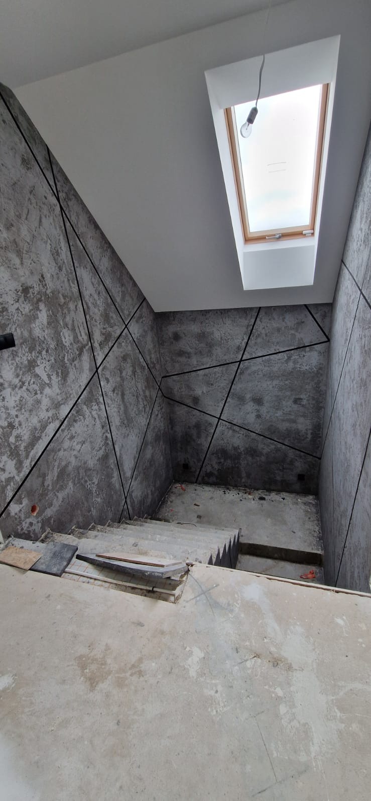 Wnętrze w trakcie prac wykończeniowych: ściany z szarą fakturą i czarnymi liniami, surowe schody, okno dachowe z widoczną żarówką. Widok z góry na dół.