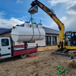 BIOLEN GRZEGORZ RYBIŃSKI - Żółta koparka Komatsu podnosi za pomocą pasów biały zbiornik na tle domu z ciemną dachówką, zbiornik umieszczany na czerwonej platformie białego samochodu dostawczego.