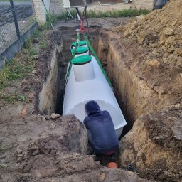 BIOLEN GRZEGORZ RYBIŃSKI - Montaż białego zbiornika z zielonymi pokrywami w głębokim wykopie; pracownik w niebieskiej bluzie przy zbiorniku, na górze łyżka koparki na pasach.