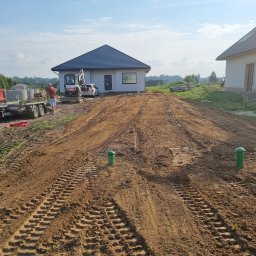 BIOLEN GRZEGORZ RYBIŃSKI - Przygotowanie terenu pod budowę domu jednorodzinnego: wyrównany teren z widocznymi śladami opon, minikoparka Bobcat i przyczepa z kręgami betonowymi w tle, zielone słupki geodezyjne na pierwszym...