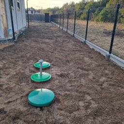 BIOLEN GRZEGORZ RYBIŃSKI - Przydomowa oczyszczalnia ścieków z trzema zielonymi włazami na gruncie, widok z góry, w tle ogrodzenie panelowe i surowy budynek w trakcie budowy.