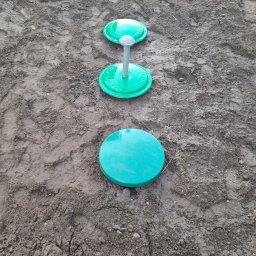 BIOLEN GRZEGORZ RYBIŃSKI - Trzy zielone pokrywy studzienek kanalizacyjnych, ułożone w linii prostej na ubitej, szarej ziemi z widocznymi śladami opon.