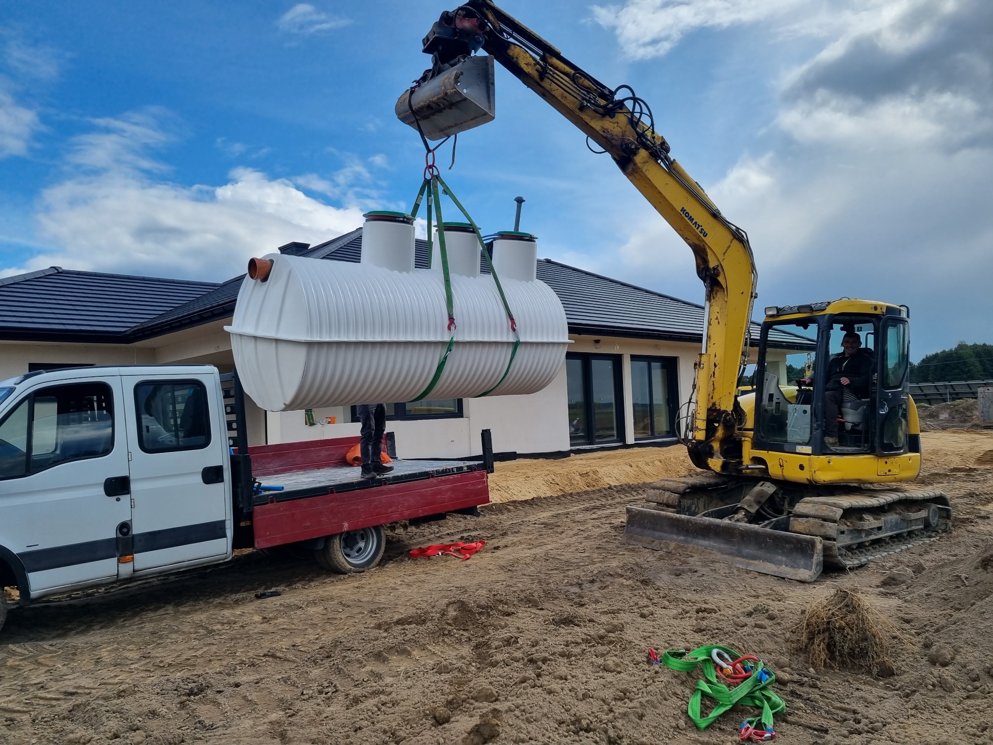 Żółta koparka Komatsu podnosi za pomocą pasów biały zbiornik na tle domu z ciemną dachówką, zbiornik umieszczany na czerwonej platformie białego samochodu dostawczego.