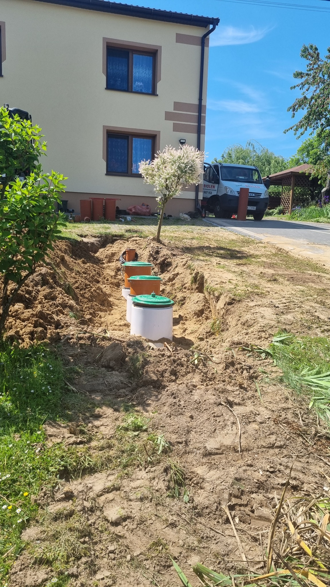 Instalacja trzech zbiorników z zielonymi pokrywami w wykopie ziemnym na tle domu jednorodzinnego i zaparkowanego białego samochodu dostawczego z reklamą firmy.