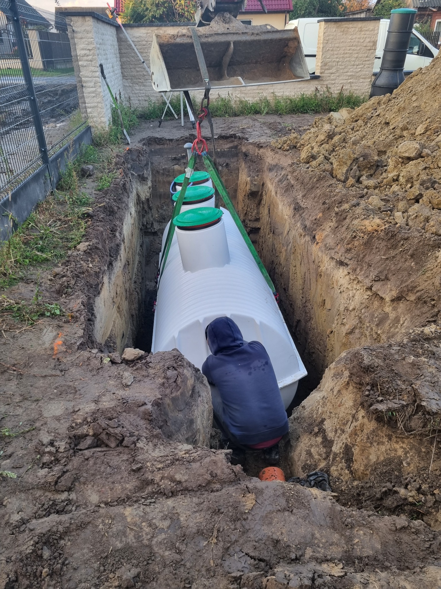 Montaż białego zbiornika z zielonymi pokrywami w głębokim wykopie; pracownik w niebieskiej bluzie przy zbiorniku, na górze łyżka koparki na pasach.