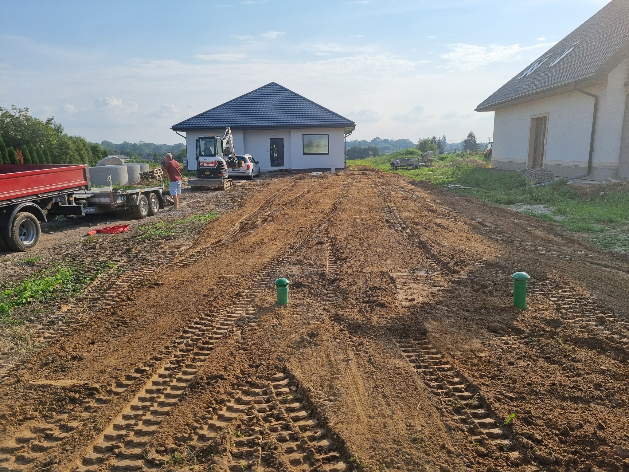 Przygotowanie terenu pod budowę domu jednorodzinnego: wyrównany teren z widocznymi śladami opon, minikoparka Bobcat i przyczepa z kręgami betonowymi w tle, zielone słupki geodezyjne na pierwszym...