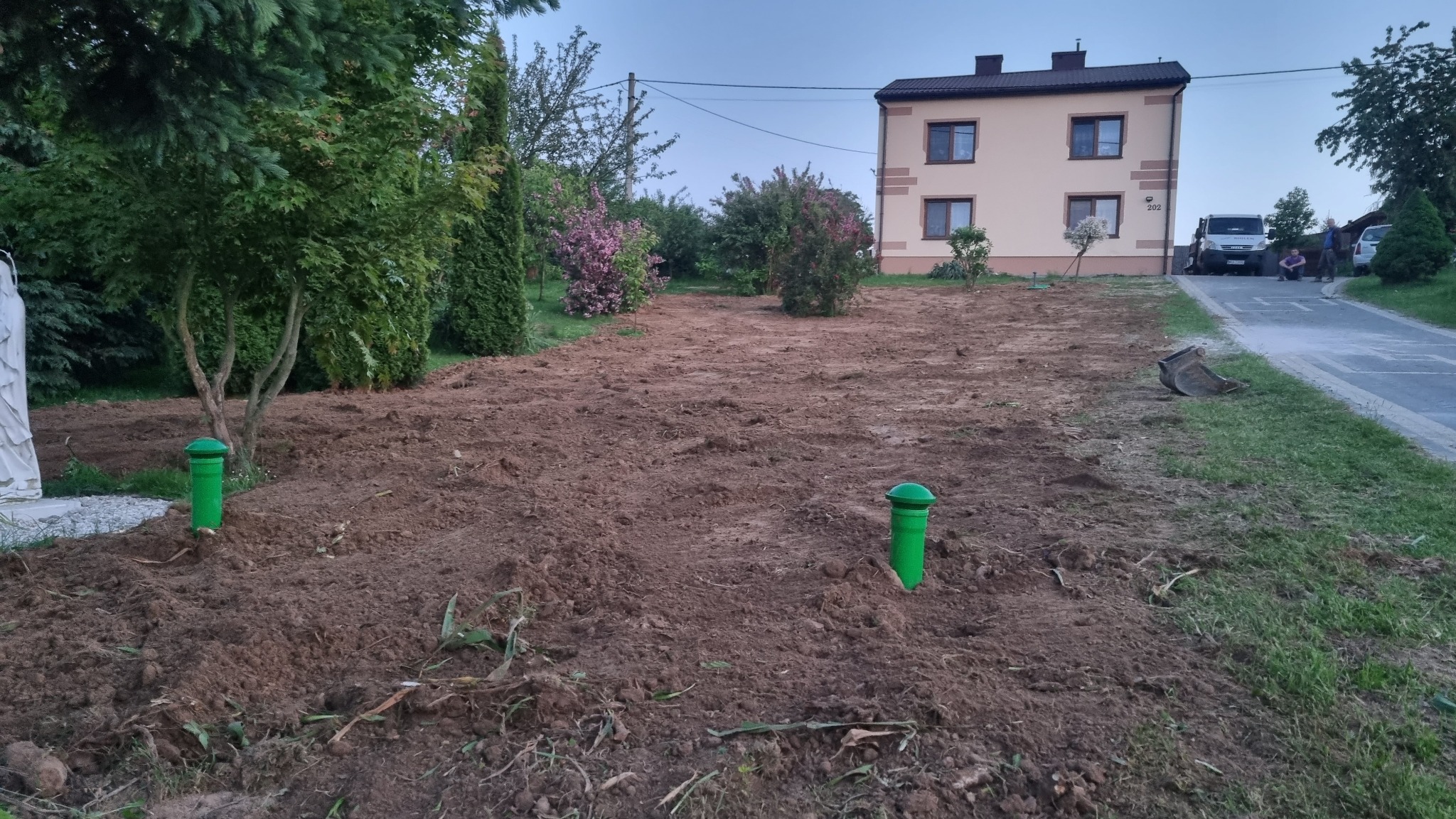 Przygotowany teren pod instalację z dwoma zielonymi odpowietrznikami, widoczny budynek mieszkalny w tle oraz częściowo usunięta warstwa ziemi.