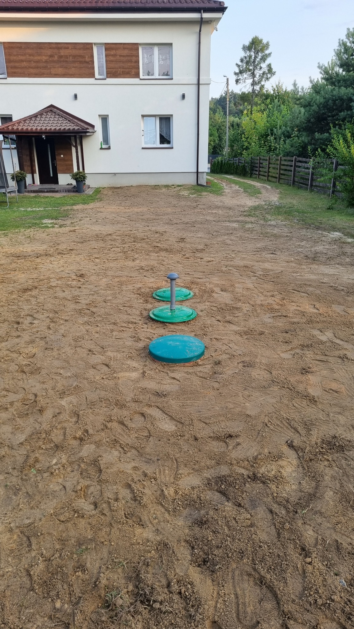 Trzy zielono-niebieskie włazy inspekcyjne ułożone w linii prostej na piaszczystym terenie przed domem jednorodzinnym.