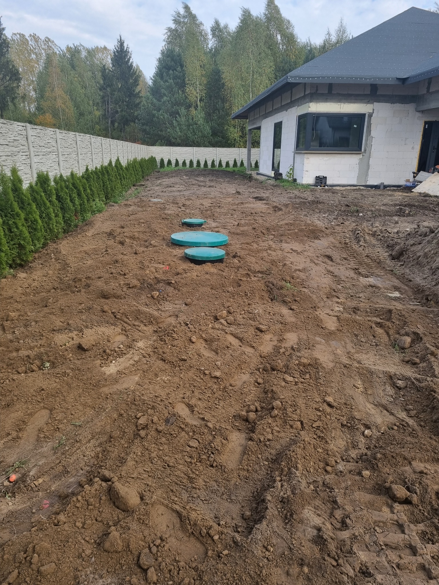 Instalacja przydomowej oczyszczalni ścieków: trzy zielone włazy na wykopanym terenie, w tle ogrodzenie betonowe z nasadzeniami iglaków i budynek w stanie surowym otwartym.
