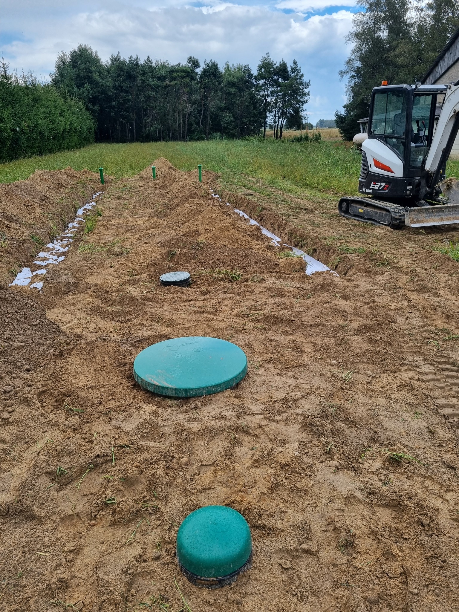 Wykopane rowy z geowłókniną, zielone kominki wentylacyjne i dwa włazy inspekcyjne na piaskowym podłożu, z mini koparką Bobcat E27Z w tle.