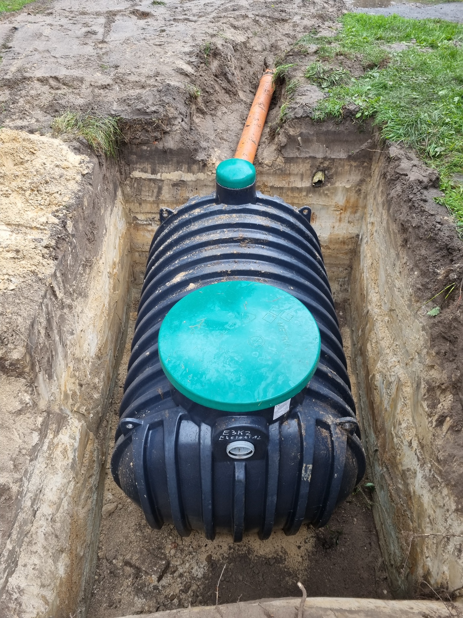 Czarny zbiornik z zielonymi pokrywami, umieszczony w wykopie z widoczną rurą odpływową, częściowo zasypany ziemią.