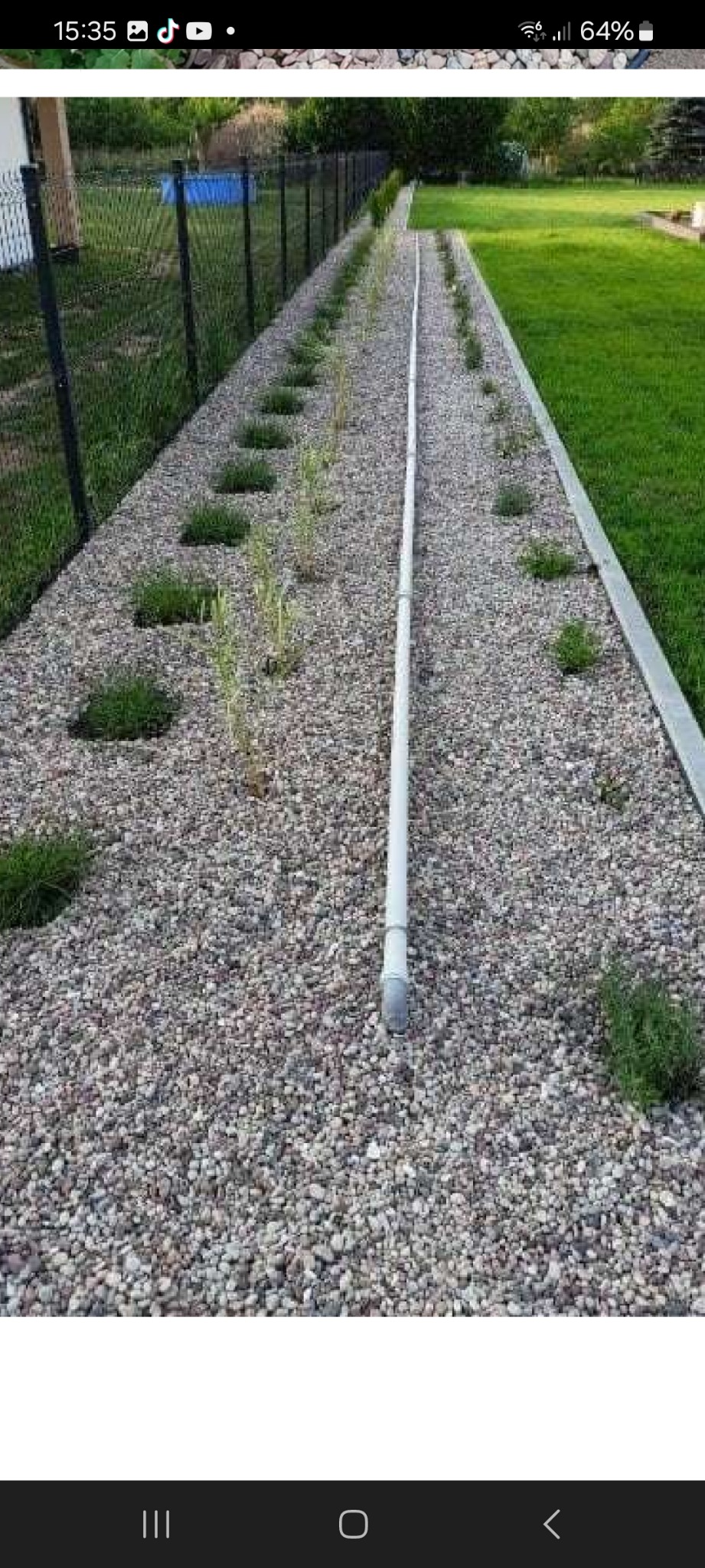 Długa, biała rura ułożona na żwirowym podłożu wzdłuż ogrodzenia z nasadzeniami, prawdopodobnie element instalacji sanitarnej.