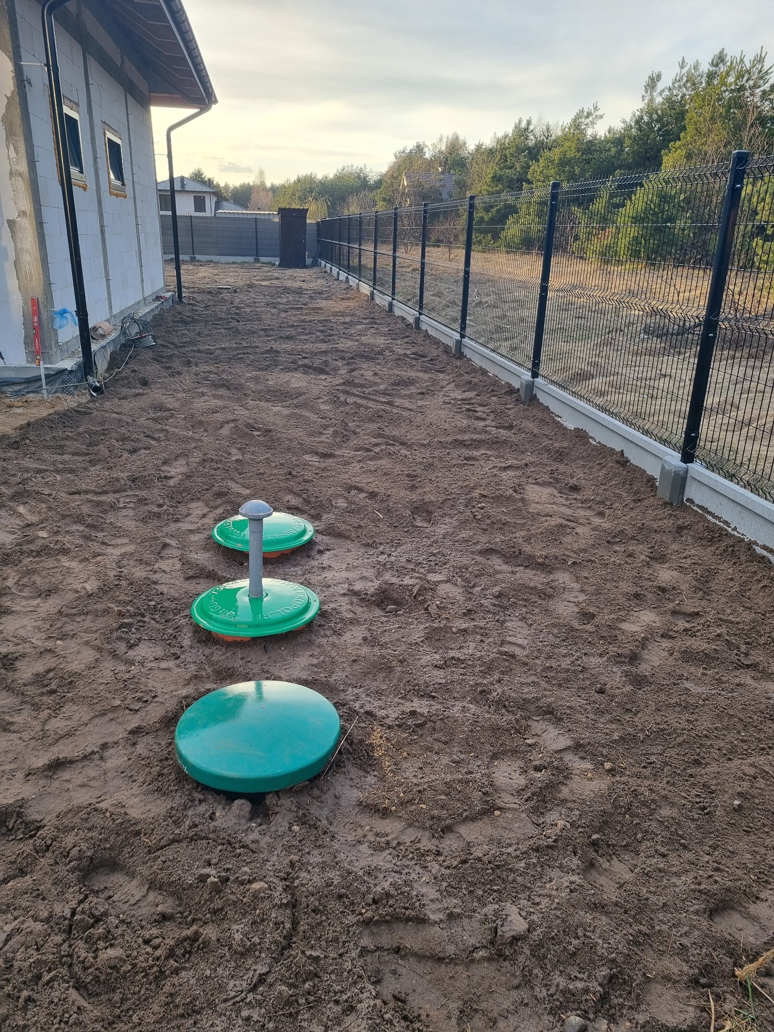 Przydomowa oczyszczalnia ścieków z trzema zielonymi włazami na gruncie, widok z góry, w tle ogrodzenie panelowe i surowy budynek w trakcie budowy.