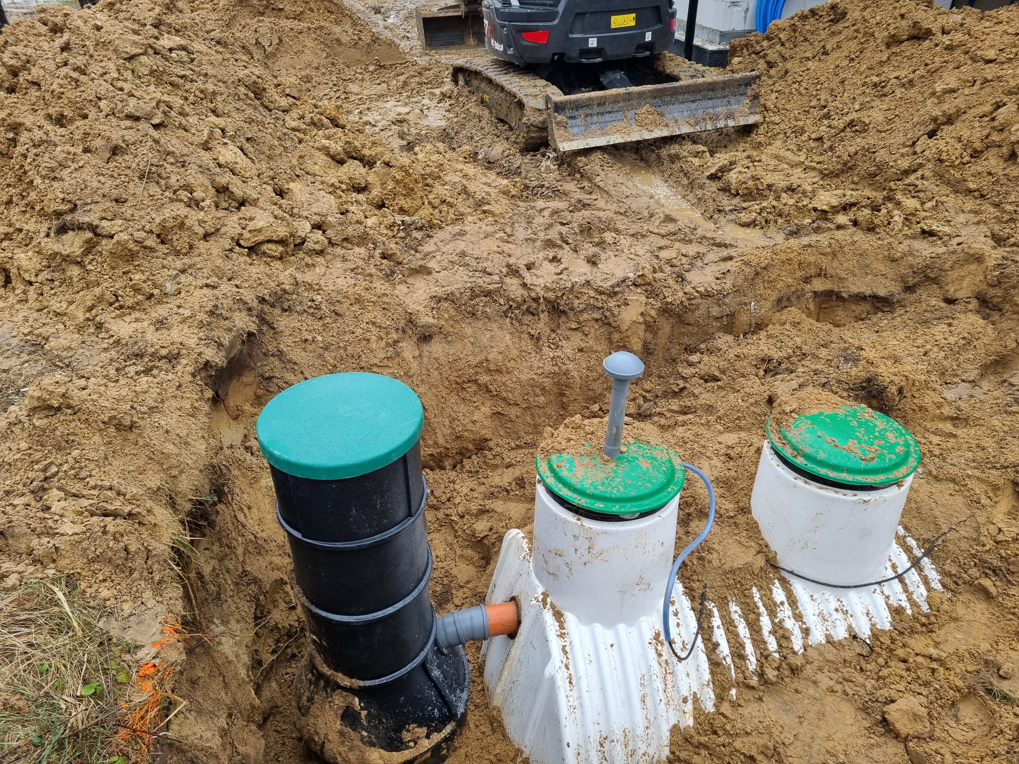 Instalacja trzech zbiorników z zielonymi pokrywami w wykopie, z widocznym mini-koparką w tle na grząskiej ziemi.