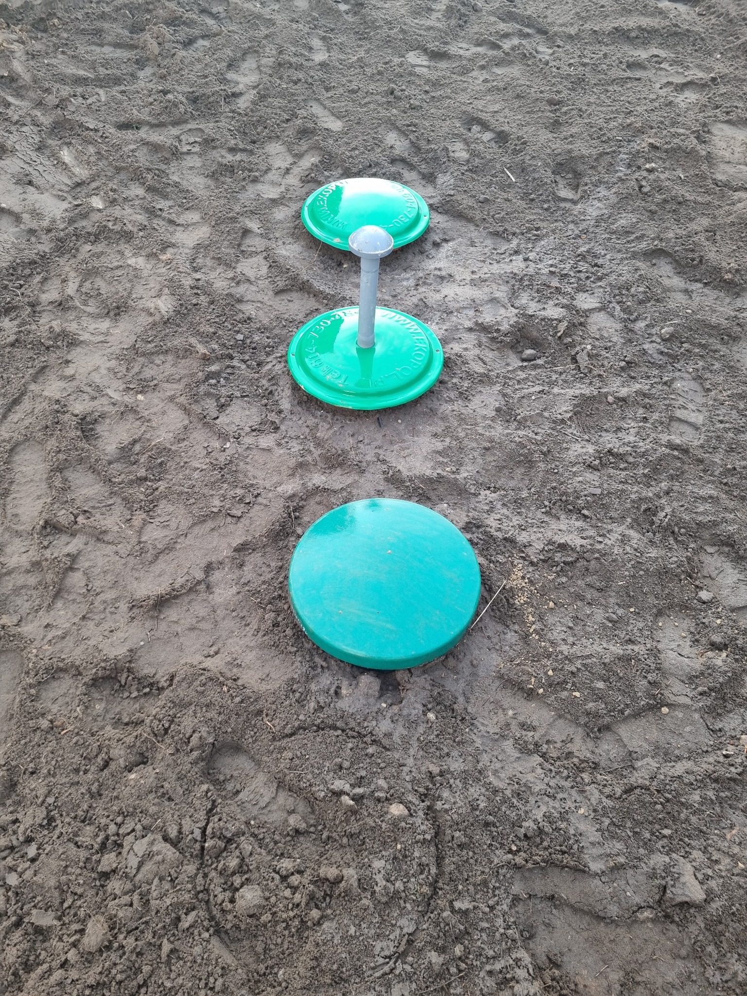 Trzy zielone pokrywy studzienek kanalizacyjnych, ułożone w linii prostej na ubitej, szarej ziemi z widocznymi śladami opon.