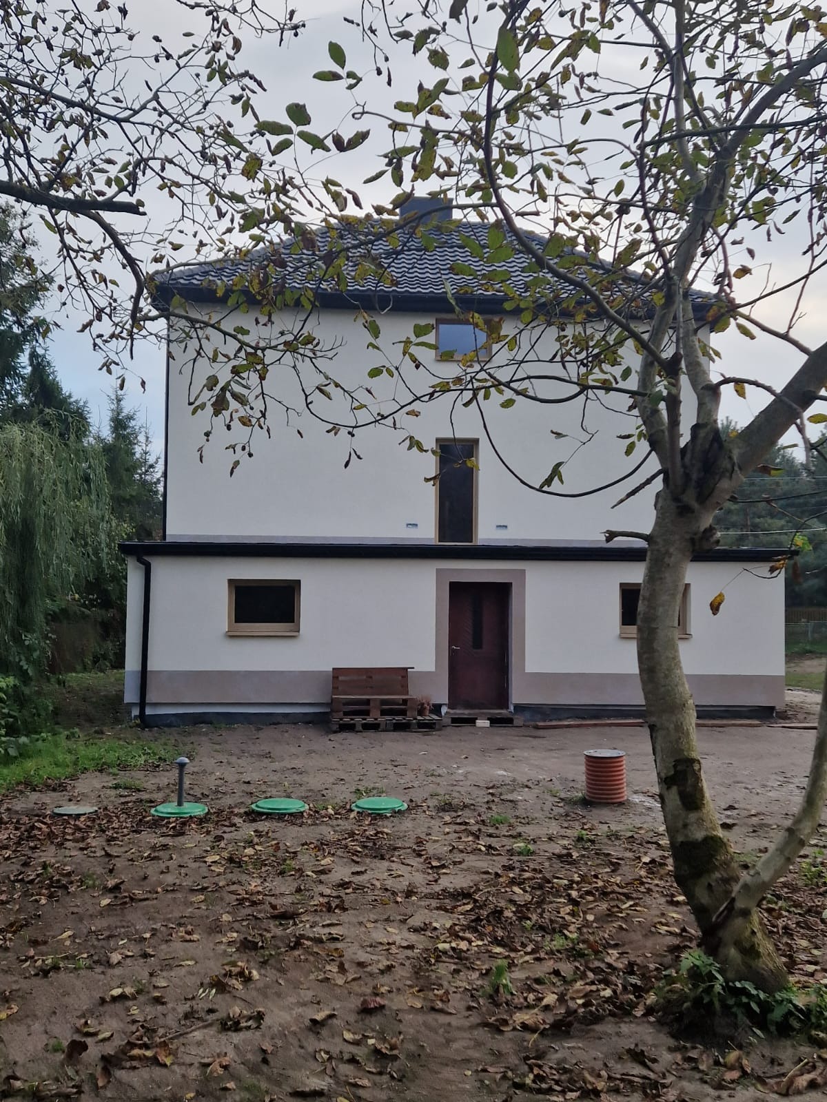 Dwukondygnacyjny dom z szarego tynku z trzema zielonymi pokrywami studzienek na pierwszym planie i paletą drewnianą przy ścianie.