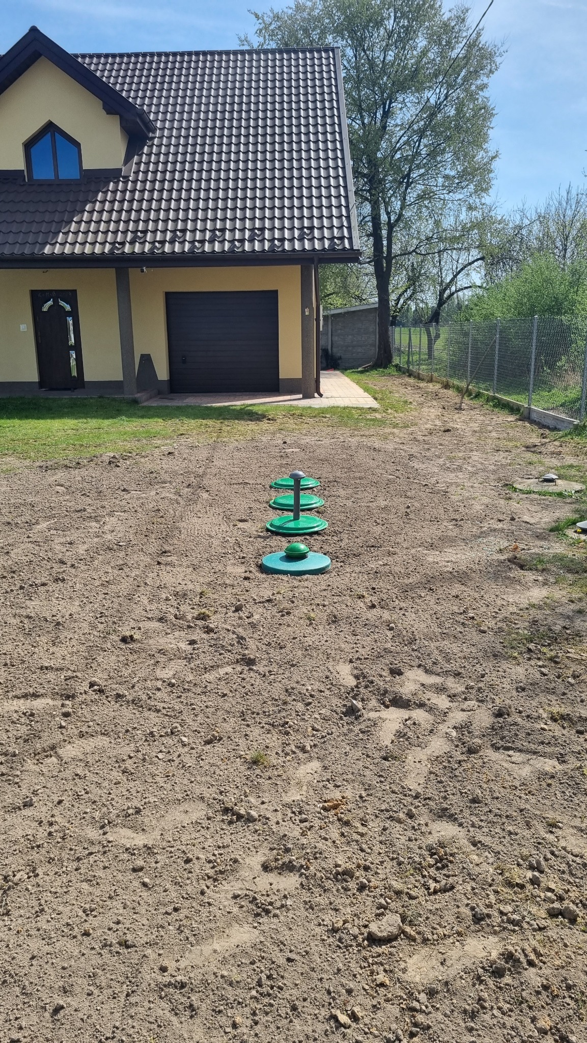 Zielone elementy wentylacyjne wystające z ziemi na przygotowanym placu budowy, dom jednorodzinny w tle.
