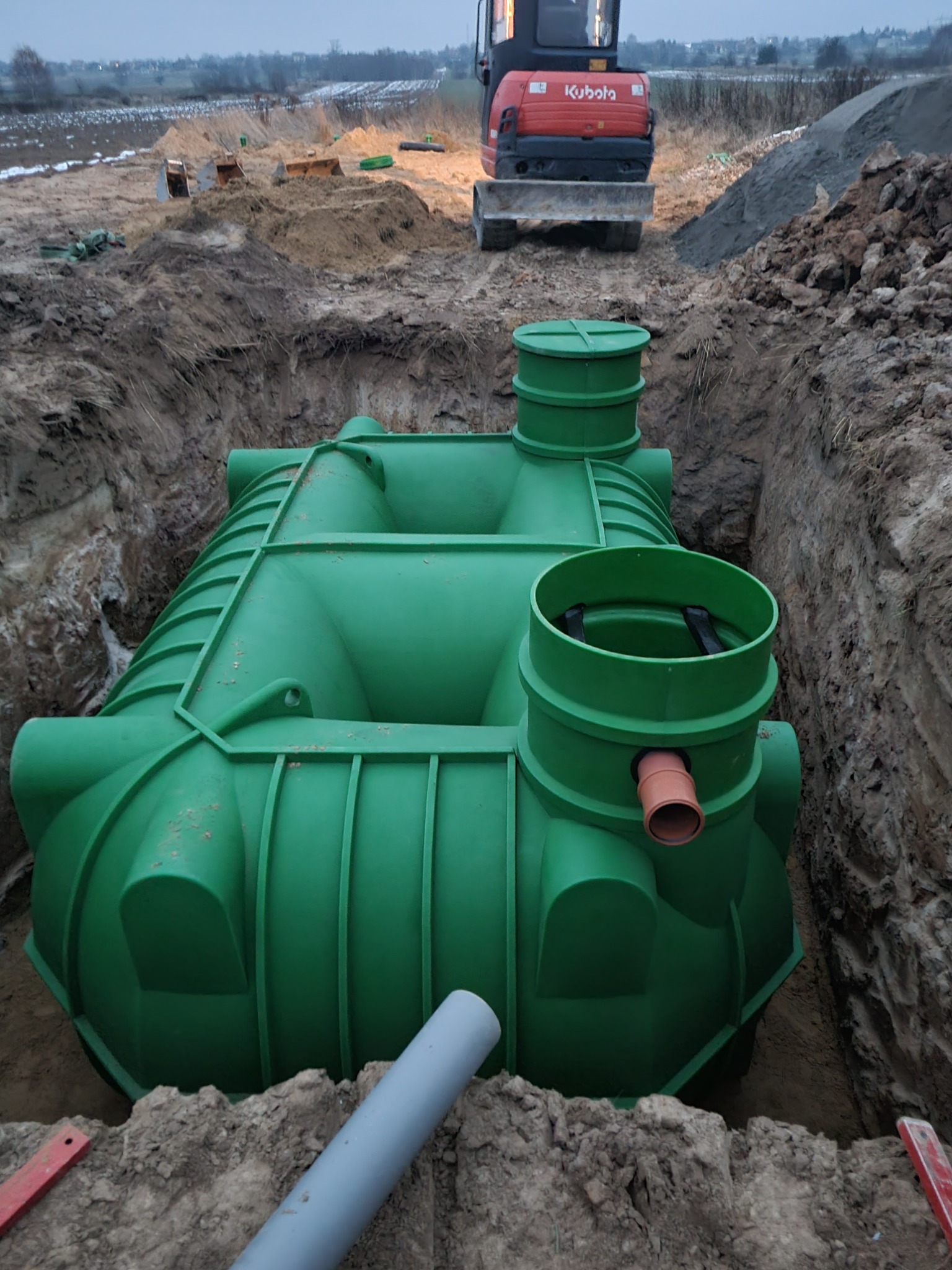 Zielony zbiornik z tworzywa sztucznego umieszczony w wykopie, widoczny fragment rury, w tle koparka Kubota i pryzma ziemi.