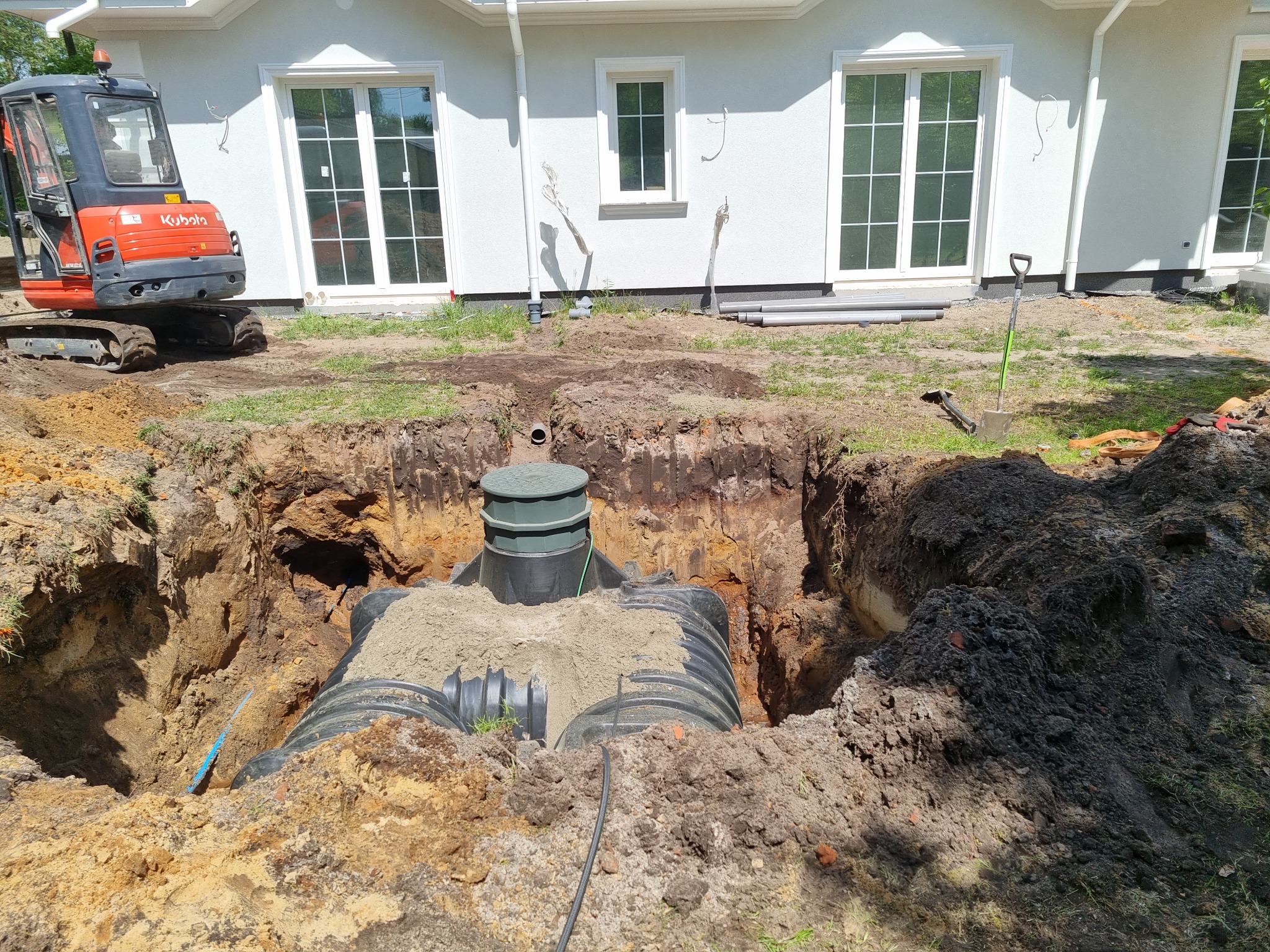 Instalacja przydomowej oczyszczalni ścieków w wykopie, widoczny zbiornik z pokrywą, zasypany piaskiem, obok mini koparka Kubota i narzędzia.