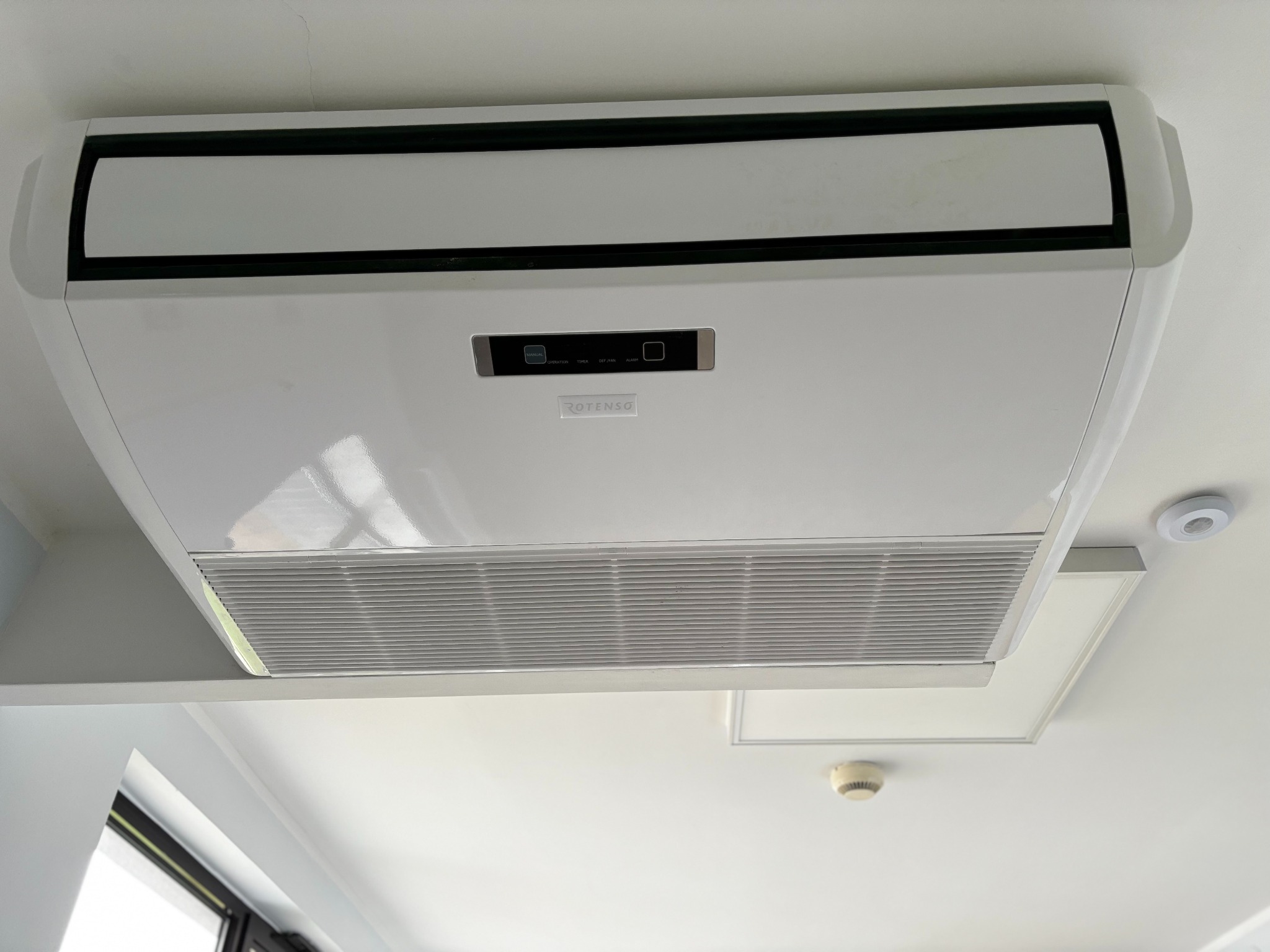 Sufitowa kaseta klimatyzacyjna marki Rotenso z widocznym panelem sterowania i kratką wylotową, zamontowana w pomieszczeniu z czujnikiem dymu i czujnikiem ruchu.