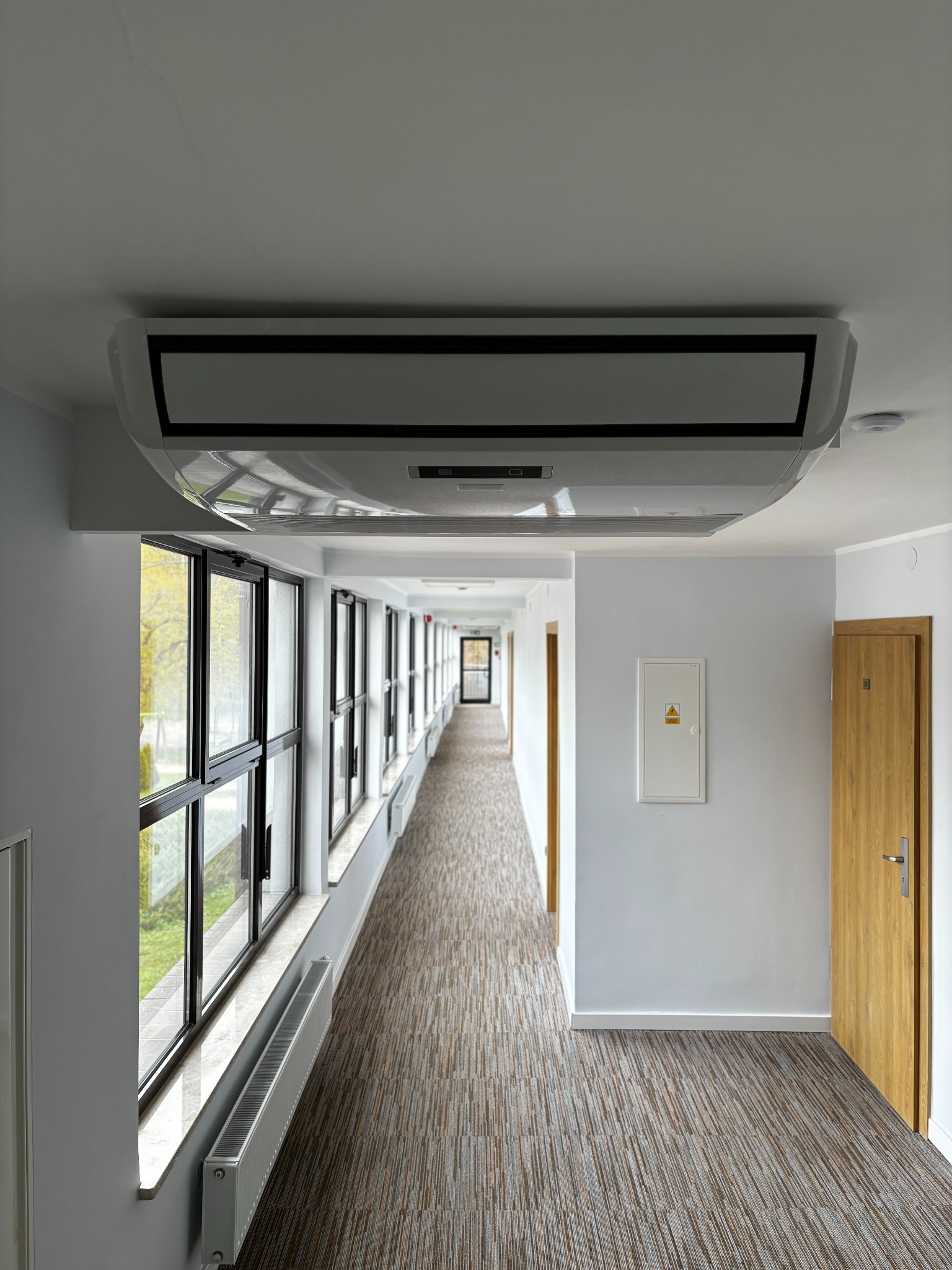Długi korytarz biurowy z oknami i grzejnikami, wyposażony w sufitowy klimakonwektor kasetonowy oraz drzwi wejściowe.