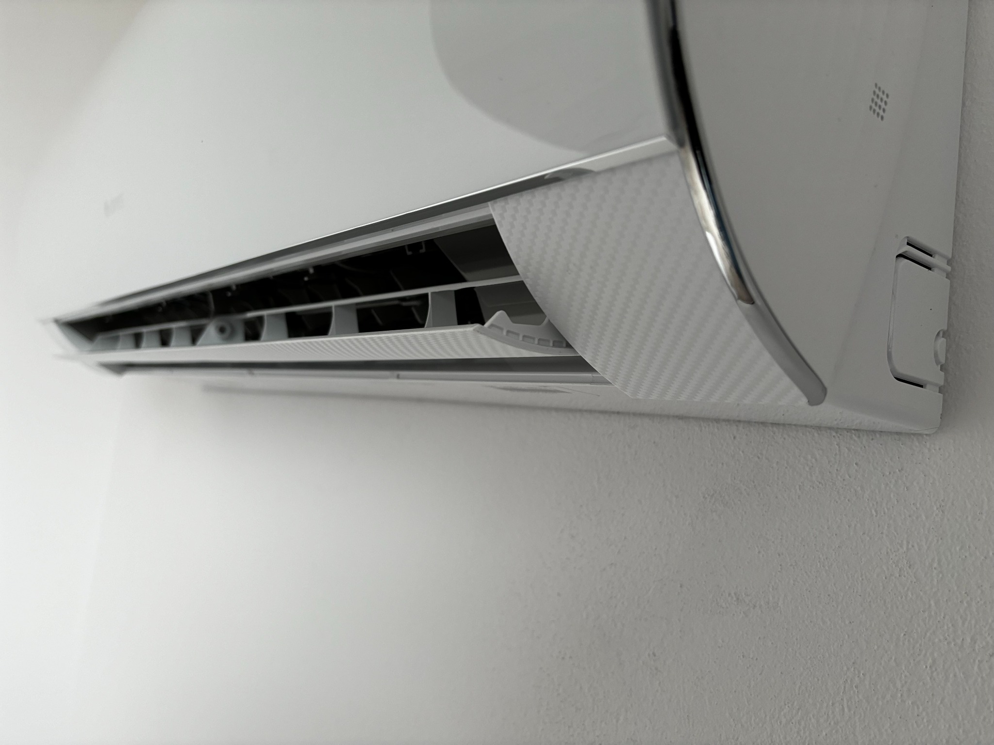 Biały klimatyzator ścienny z widocznymi żaluzjami wylotu powietrza, zamontowany na białej, teksturowanej ścianie.