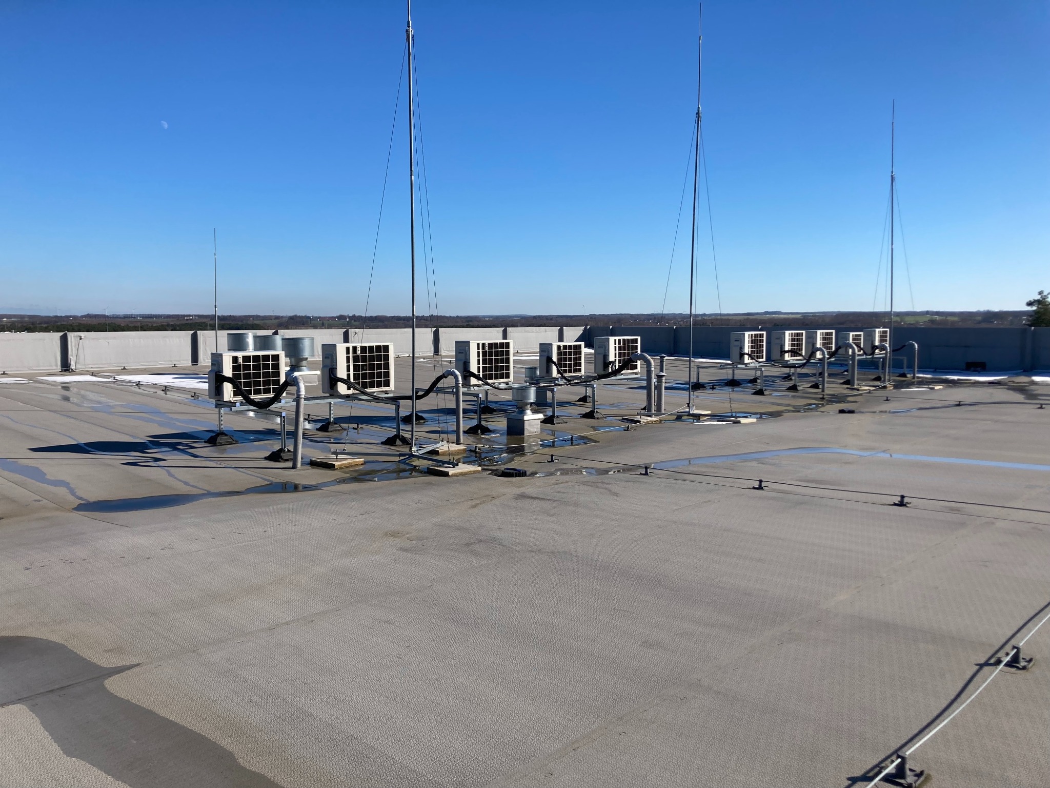 Rząd klimatyzatorów na płaskim dachu budynku komercyjnego z widocznymi odgromnikami i niewielkimi kałużami wody.