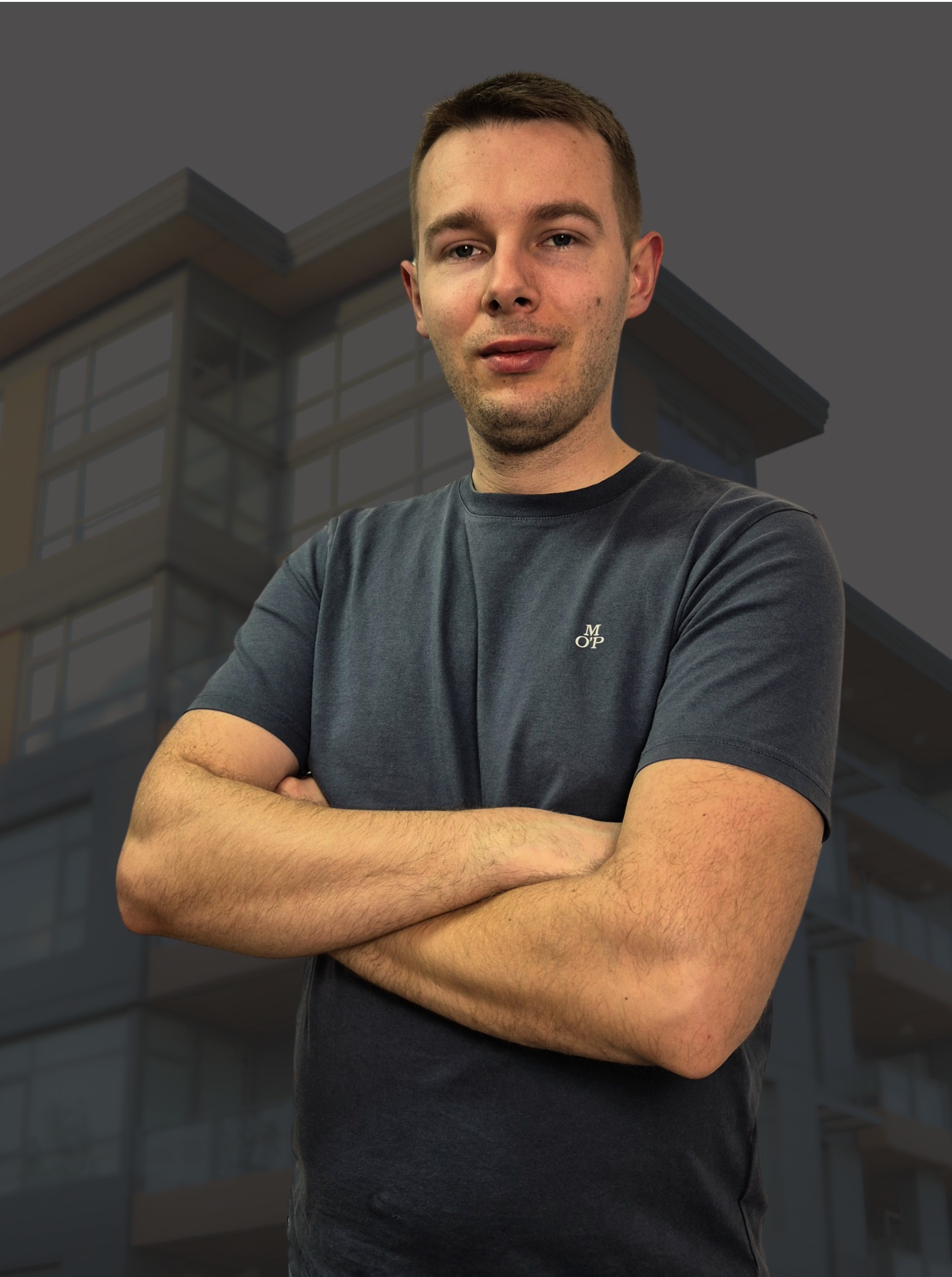 Mężczyzna w szarej koszulce z założonymi rękami na tle budynku mieszkalnego o nowoczesnej architekturze.