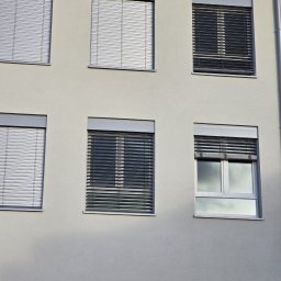 ZPHU BARTEK - Fasada budynku z sześcioma oknami, z których część posiada zewnętrzne osłony okienne z regulowanymi lamelami, ustawionymi pod różnymi kątami, wpływając na stopień zaciemnienia wnętrza.