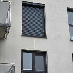 ZPHU BARTEK - Szara fasada budynku z zamkniętą, poziomą osłoną okienną w kolorze antracytowym oraz oknem z zamontowaną moskitierą.