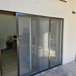 ZPHU BARTEK - Przesuwne drzwi tarasowe z moskitierą w nowoczesnym domu, widok z zewnątrz na taras wyłożony kostką brukową, w tle las.