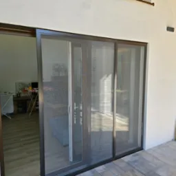 Przesuwne drzwi tarasowe z moskitierą w nowoczesnym domu, widok z zewnątrz na taras wyłożony kostką brukową, w tle las.