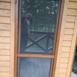 ZPHU BARTEK - Drewniane drzwi z moskitierą, widoczne przez siatkę drzewa i fragment balustrady balkonu, odbicie osoby w siatce.