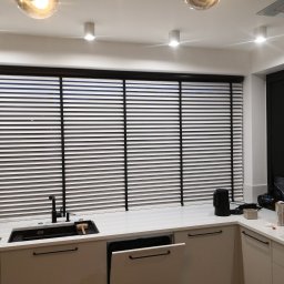 ZPHU BARTEK - Kuchnia z białymi szafkami i czarnymi żaluzjami drewnianymi, widoczny zlew z czarną armaturą i akcesoria kuchenne na blacie.