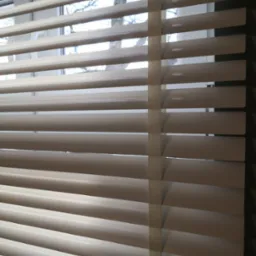 Beżowe poziome listwy osłonowe okna z widocznym drzewem za szybą, regulowane sznurkiem po prawej stronie.