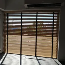 Jasne, drewniane lamele z czarnymi taśmami na oknie, częściowo otwarte, wpadające promienie słoneczne tworzą cień na parapecie, widok na budynki i zieleń za oknem.