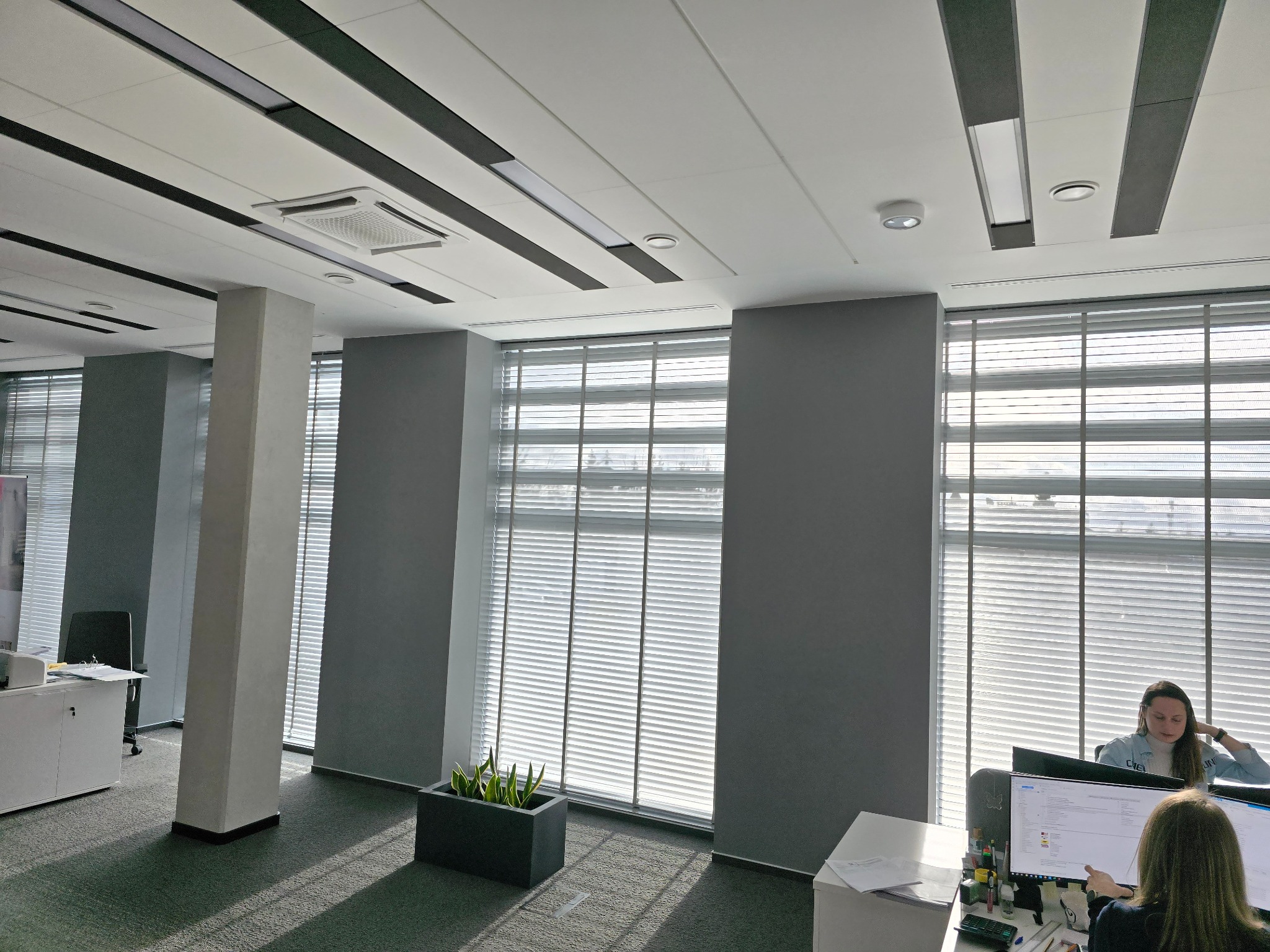 Biuro z aluminiowymi żaluzjami poziomymi w oknach, widoczne stanowiska pracy z pracownikami.